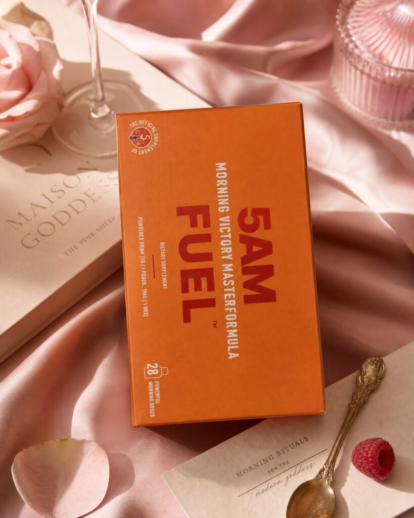 A box of 5AM FUEL morning drink sachets on pink silk beside a Maison Goddess notebook